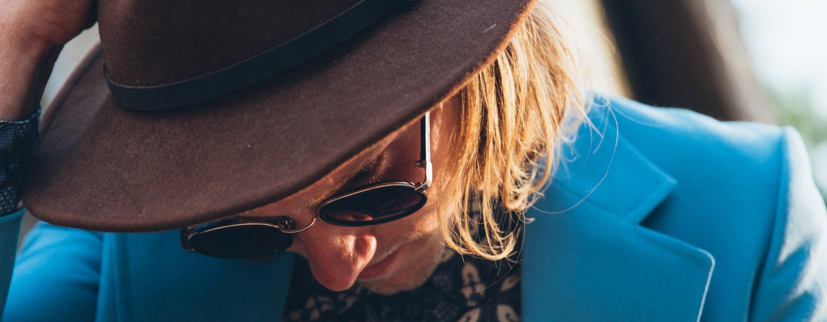 Person wearing a brown hat and blue blazer with sunglasses, leaning against a wall.